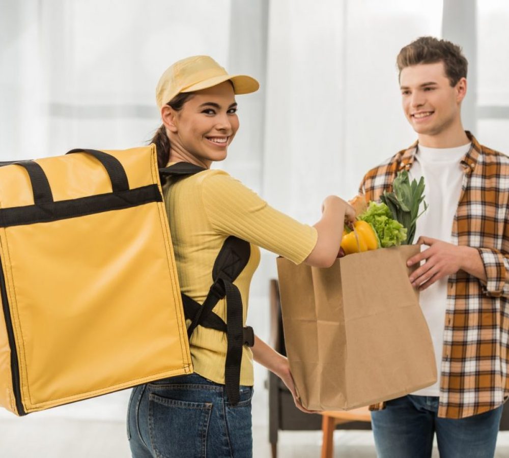 attractive-courier-smiling-at-camera-and-giving-package-with-fresh-vegetables-to-man-at-home.jpg